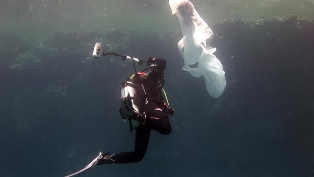 Young woman underwater model in white cloth and cameraman. Creativity and art of creating images on background of seabed.