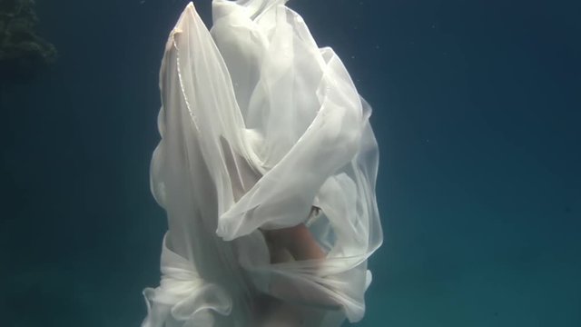 Young woman underwater model in white cloth depicts birth of an angel. Creativity and art of creating images on background of seabed.