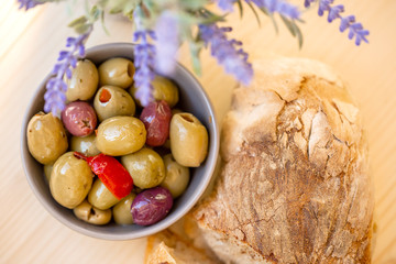 Olives stuffed with red pepper and homemade bread. Multicolored olives in small bowl on a wooden table. Lavender. Blurry background. Closeup. Soft focus. Top view.