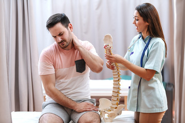 Man in pain sitting on hospital bed and holding neck. Next to him standing doctor and holding model...