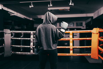 Fototapeta na wymiar Boxer girl with hoodie and boxing gloves on standing in ring with backs turned. One hand lifted up.