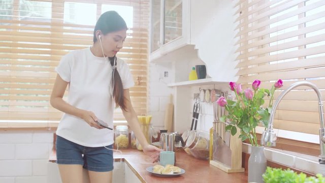 Young Asian teenager woman drinking warm coffee using smartphone listening music and checking social in kitchen at home. Lifestyle woman relax in morning at home concept.
