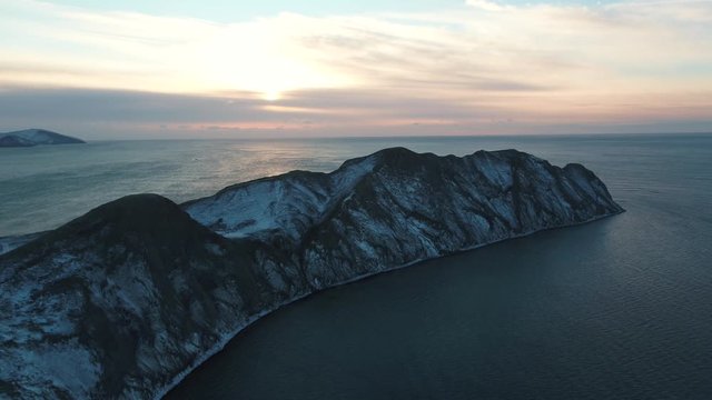 Aerial view on a rocky cape covered by snow in the dark sea water against cloudy sky at sunrise. Shot. Beautiful marine landscape.