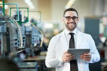 Waist up portrait of handsome factory worker holding digital tablet posing  in workshop and smiling...