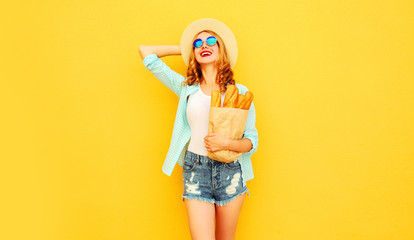 portrait happy smiling young woman holding paper bag with long white bread baguette, wearing straw hat, shorts on colorful yellow background