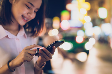 Closeup of woman holding a smartphone at night on city shopping street, and people walk. Technology for communication concept.