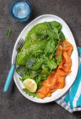 Low carbs salad. Spinach, rucola salad with avocado and salmon. Black concrete background, white plate, top view