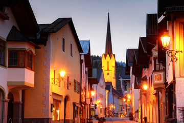Narrow main street in blue hour in Bavarian resort town Garmisch-Partenkirchen historic center with tall spire of Maria Himmelfahrt church above row of traditional German houses, Bayern Germany Europe