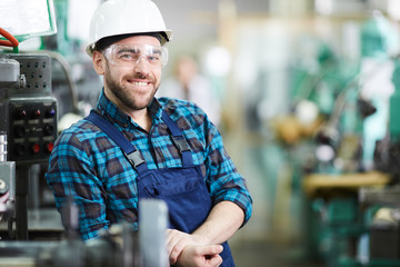 Waist up portrait of bearded factory worker wearing hardhat smiling at camera while posing in...