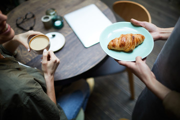 Above view of unrecognizable waitress bringing crispy croissant on plate to customer drinking tasty coffee in cafe