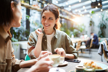 Positive excited young women in casual clothing sitting at table and looking at each other while discussing last news and drinking coffee in cafe