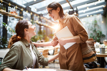 Excited surprised young lady in glasses holding laptop and touching shoulder of friend while greeting her in cafe, girl bumping into friend in public place