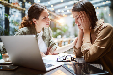 Sad young woman puzzled by problems sitting at table with laptop, tablet and papers and leaning head on hands while thinking about her failure, her friend supporting her in difficult situation