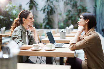 Positive confident business ladies in casual shirts sitting at table in cafe and discussing project and plans, they using laptop