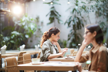 Serious busy young lady with curly hair sitting at table in cafe with eco design and using laptop...