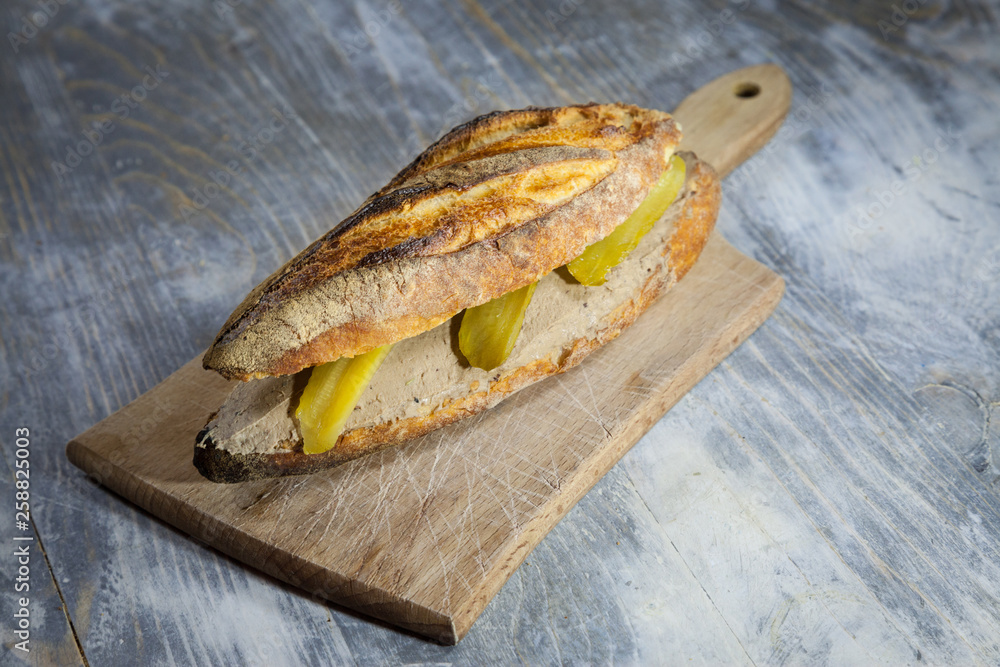 Wall mural Liver Pate sandwich in a French baguette, made of duck and chicken Pate de Foie with some sliced pickles, on display on a rustic wooden table. It is a typical French snack