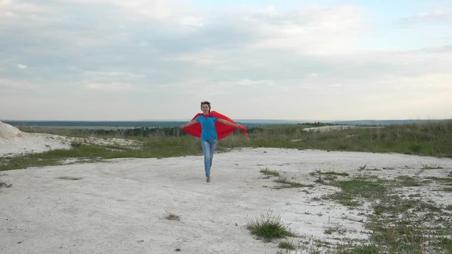 happy girl superhero runs across field in red cloak in front of blue sky and smiles. slow motion. young girl dreams of becoming superhero. happy teen girl goes in red cloak expression of dreams.