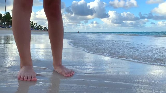 Young woman standing barefoot on the tropical sandy beach and splashed by ocean waves. Cloudy skies and blue ocean waves.