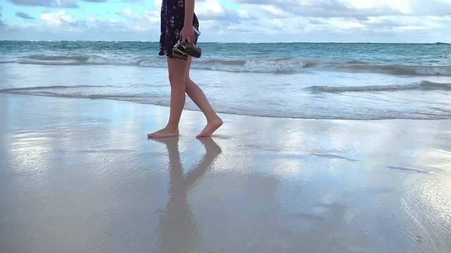Young woman in summer dress walking barefoot on beautiful tropical beach, and feet soaked by the ocean waves.