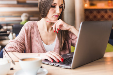 Attractive young woman sits at a table in a cafe with a cup of coffee and enjoys a laptop