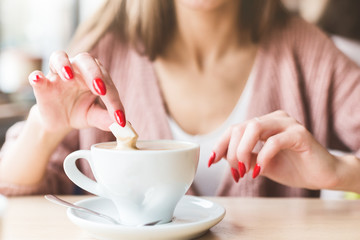 A girl dunks a piece of sugar in coffee. Closeup of hands with red nails make-up and cappuccino cup