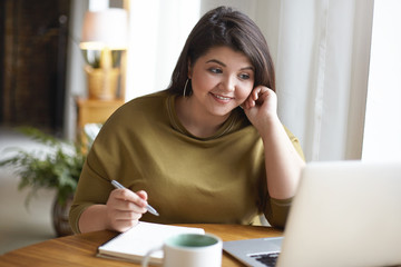 Indoor image of cheerful overweight student girl working on course paper, usign wireless internet connection on generic portable computer, making notes in copybook and smiling. Gadgets and education - obrazy, fototapety, plakaty