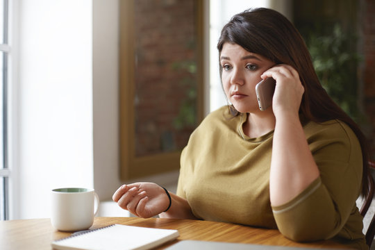 Picture of serious young plus size Caucasian woman speaking on smart phone sitting at cafe table in front of open copybook and mug, having worried look, upset with negative news. Selective focus