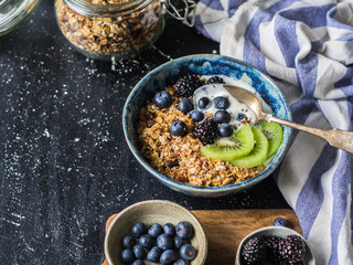 Healthy breakfast - homemade granola with yogurt, berries, fruit in blue bowl on dark background. 