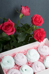Homemade marshmallows of different colors are beautifully packed in a gift box. Nearby is the lid of the box with a transparent window. Nearby is a bouquet of scarlet roses.