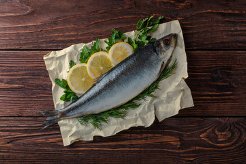 Herring lies on paper with lemon slices, onion rings, parsley, dill. Salted herring lies on a brown wooden table. View from above.