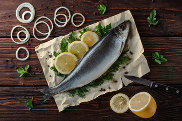 Herring lies on paper with lemon slices, onion rings, parsley, dill. Salted herring lies on a brown wooden table. View from above.