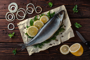 Herring lies on paper with lemon slices, onion rings, parsley, dill. Salted herring lies on a brown wooden table. View from above.