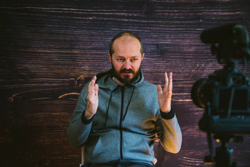 Bearded middle age man in hoody sitting and talking in front of DSLR camera, video blogging concept