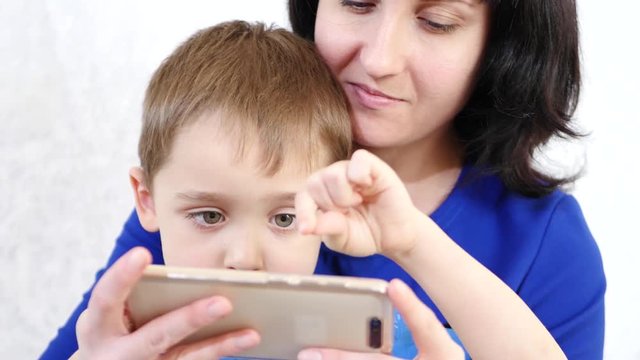 A woman holds a little boy hugging him. The child touches the touch screen of the phone and plays a child s game on a smart phone. Life is online. Education and development.