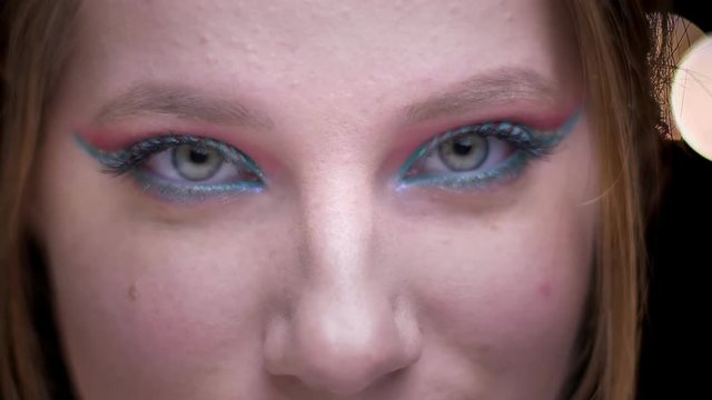 Eye-portrait of blonde model with colorful make-up raising her eyebrows and ogling the camera on blurred lights background.