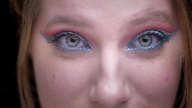 Close-up portrait of fair-haired model with colorful make-up moving her eyes back and forth on blurred lights background.