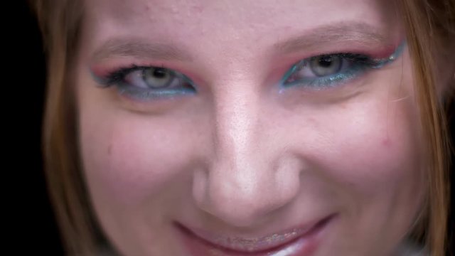 Close-up portrait of fair-haired model with colorful make-up joyfully moving her eyes on blurred lights background.
