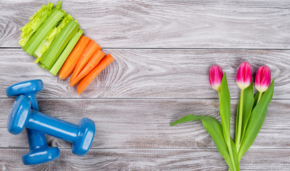 Spring flatlay sports composition with celery an carrots blue dumbbells and tulips on gray wooden...
