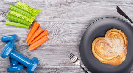 Spring flatlay sports composition with celery an carrots blue dumbbells and plate with bakery on wooden background. Concept healthy lifestyle or fastfood, sport and diet in spring. Top view.