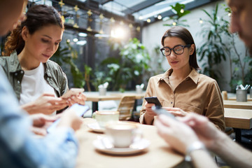 Gathering of gadget-addicted friends: serious concentrated young people sitting at table in cafe and using modern smartphones while texting messages or surfing net