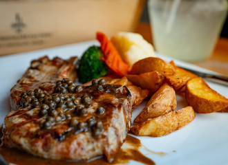 Selective focus on steak topped with sauce and pepper serve with potato fried and carrots, cauliflower and broccoli steamed. Steak on white plate put on wooden table. Tomahawk steak.