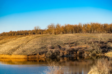 Early spring landscape, in the Rostov region in the city of Shakhty, on the Grushevka river. Sunny sunset in yellow and orange warm light. Dry grass on the local shore and a beautiful reflection in th