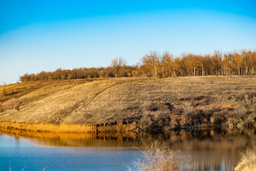 Early spring landscape, in the Rostov region in the city of Shakhty, on the Grushevka river. Sunny sunset in yellow and orange warm light. Dry grass on the local shore and a beautiful reflection in th