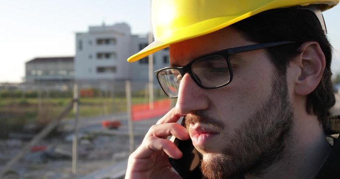 Ragazzo giovane architetto parla al telefono per affari di lavoro mentre &egrave; in cantiere con il casco protettivo giallo. Occhiali da vista, giacca colore nero e barba lunga. 