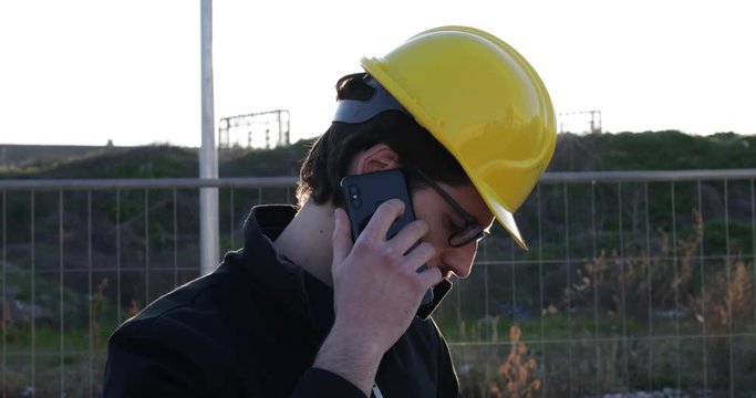Ragazzo giovane architetto parla al telefono per affari di lavoro mentre &egrave; in cantiere con il casco protettivo giallo. Occhiali da vista, giacca colore nero e barba lunga. 