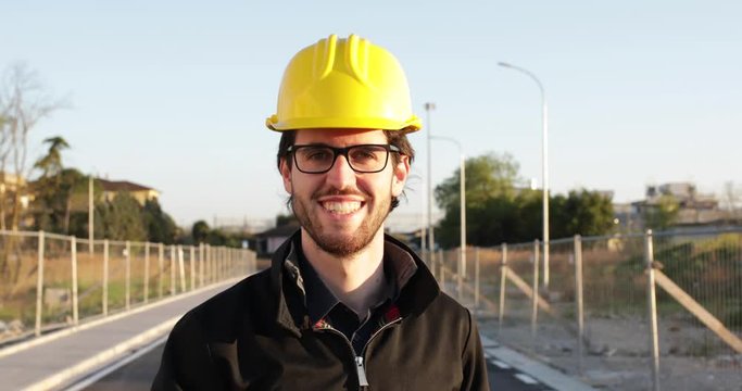 Ingegnere giovane con occhiali neri, caschetto giallo e giacca che sorride in una strada cantiere prima di andare a lavoro e progettare la futura costruzione.