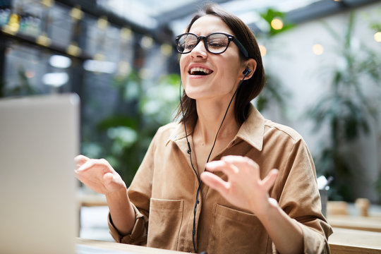 Positive carefree young woman in wired earphones sitting at table and using laptop while gesturing hands and singing favorite song at workplace