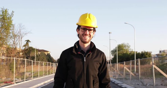 Ingegnere giovane con occhiali neri, caschetto giallo e giacca che sorride in una strada cantiere prima di andare a lavoro e progettare la futura costruzione.