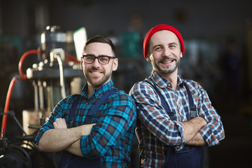 Waist up portrait of two factory workers posing in workshop standing back to back with arms...