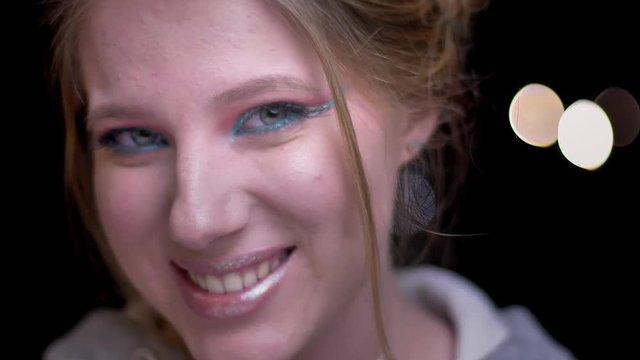 Close-up portrait of blonde girl with colorful make-up showing positive shock childishly on blurred lights background.
